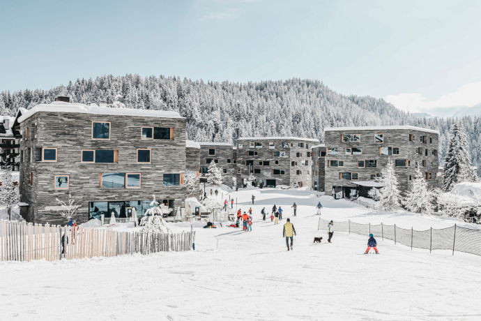 Direkt an der Talstation gelegen: Im rocksresort in Laax beginnt das Skivergnügen gleich vor der Haustür – modernes Design trifft auf alpines Lebensgefühl (Foto: © LAAX / Weisse Arena Gruppe) Winterliche Ansicht des rocksresort in Laax mit Skifahrenden vor moderner Architektur und verschneitem Wald