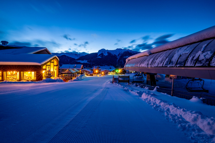 Direkt an der Talstation gelegen: Vom Boutique Hotel Pellas in Vella geht’s morgens auf die Piste und abends in die gemütliche Stube mit Blick auf die Bergwelt (Foto: © Hotel Pellas AG) Abendaufnahme des Boutique Hotel Pellas in Vella mit beleuchteter Fassade und Skipiste im Vordergrund