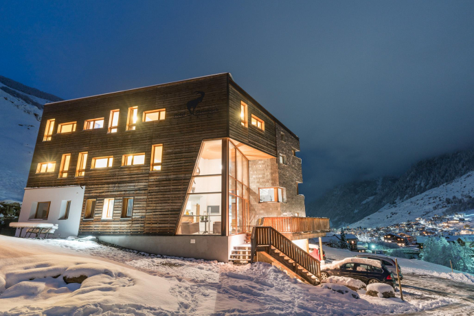Authentisch, modern, nah an der Natur: Das Hotel Steinbock in Vals besticht durch seine Holzarchitektur und die ruhige Lage oberhalb des Dorfs (Foto: © Hotel Steinbock) Hotel Steinbock in Vals bei Nacht mit beleuchteten Fenstern und verschneiter Landschaft im Hintergrund