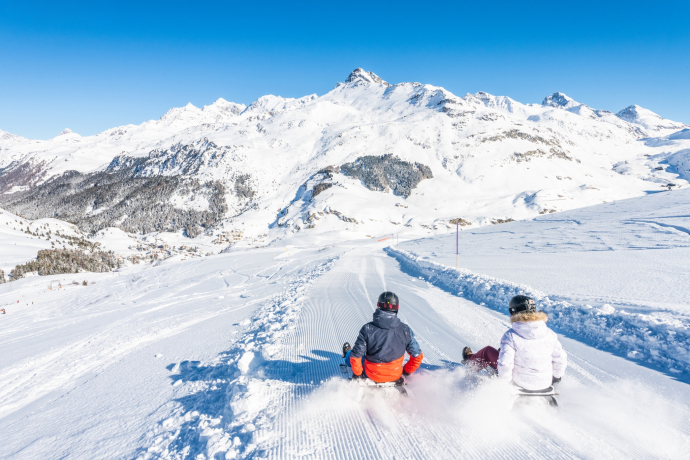 Zwei Personen fahren mit Schlitten auf einer Schlittelpiste in Bivio hinunter. Umgeben sind sie von verschneiten Bergen und klarem blauem Himmel.