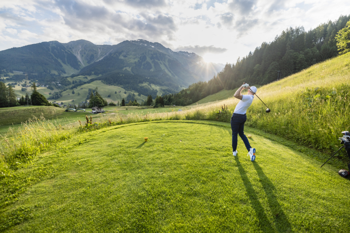 Abschlagszene auf dem Golfplatz Klosters mit sanften Hügeln, mächtigen Bergen und warmem Abendlicht im Hintergrund.