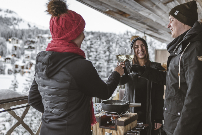 Ein Trio geniesst auf einem verschneiten Hotelbalkon ein warmes Fondue und stösst mit Weisswein an, umgeben von Winterlandschaft und gemütlicher Atmosphäre.