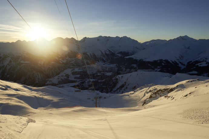 Frisch präparierte Skipiste in Disentis Sedrun, umgeben von tief verschneiten Bergen, während die Sonne über dem Gipfel erscheint.