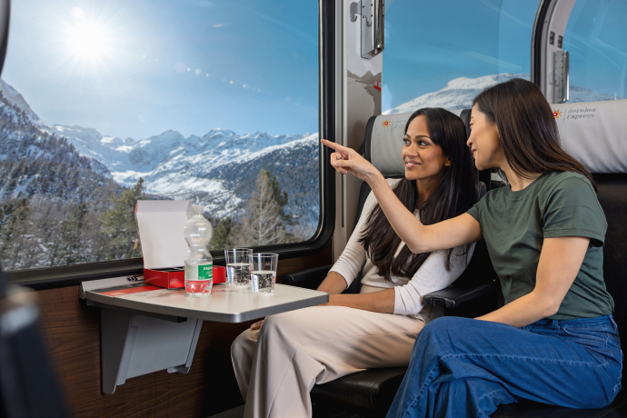 Zwei Reisende im Bernina Express schauen durch das Panoramafenster auf die verschneite Berglandschaft in Graubünden
