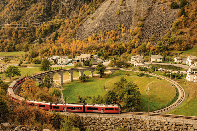 Zug der Rhätischen Bahn verlässt im Herbst das Kreisviadukt von Brusio, umgeben von herbstlich gefärbten Bäumen