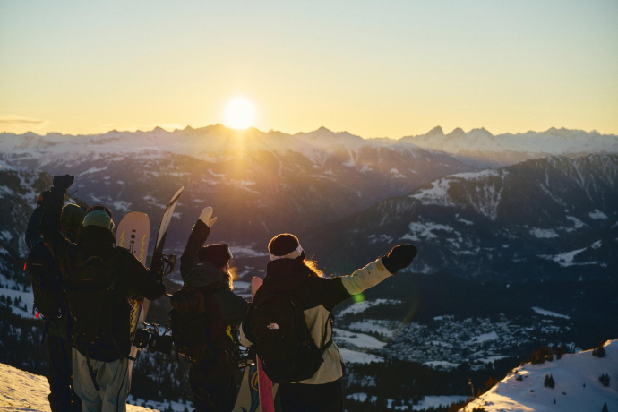 Wintersportler begrüssen den Sonnenaufgang am Gipfel, mit Blick auf verschneite Berge.