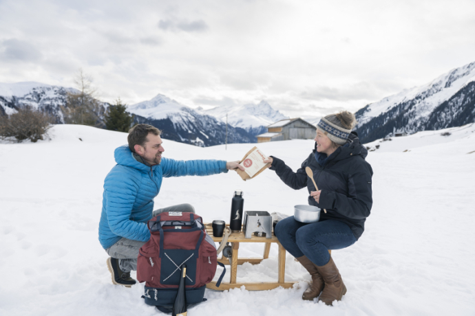 Paar bereitet Fondue im Fondue-Schlitten in Tenna zu und verschneiten Bergen im Hintergrund.