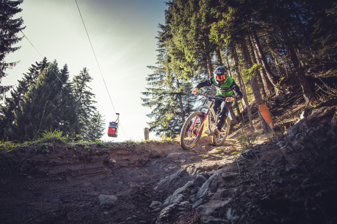 Mountainbiker auf einem bewaldeten Abschnitt im Alpenbikepark Chur mit einer vorbeifahrenden Gondel im Hintergrund