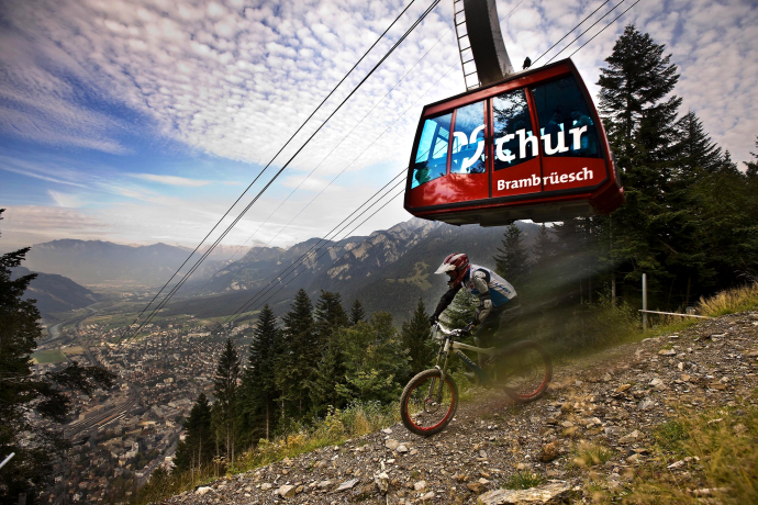 Mountainbiker fährt unter der roten Brambrüesch-Gondelbahn im Alpenbikepark Chur mit Blick auf das Rheintal