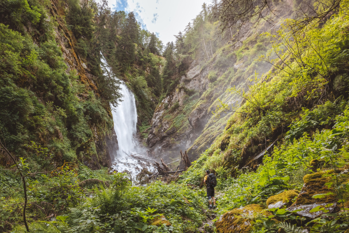 Ein Wanderer mit Rucksack steht auf einem Pfad inmitten von Farnen und blickt auf die herabstürzende Cascada da Pisch im Val Müstair