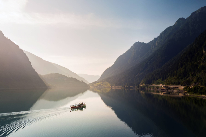 Ein kleines Schiff fährt über den ruhigen Lago di Poschiavo. Die umliegenden bewaldeten Berghänge spiegeln sich im Wasser.