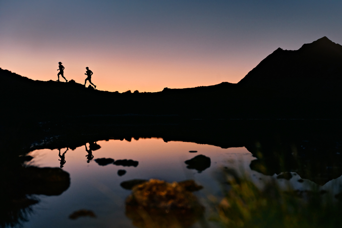 Silhouetten von zwei Trailrunnern spiegeln sich in einem Bergsee in Samnaun bei Sonnenuntergang