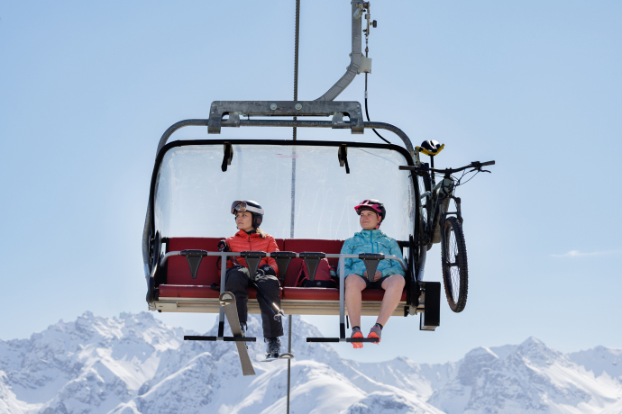 Eine Skifahrerin mit Skiausrüstung und eine Mountainbikerin mit Velo und Helm nebeneinander auf einem Sessellift vor schneebedeckten Bergen