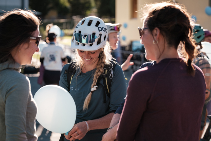 Lächelnde Mountainbikerin mit weissem Helm, Zopf und hellblauem Luftballon im Gespräch mit zwei weiteren Teilnehmerinnen