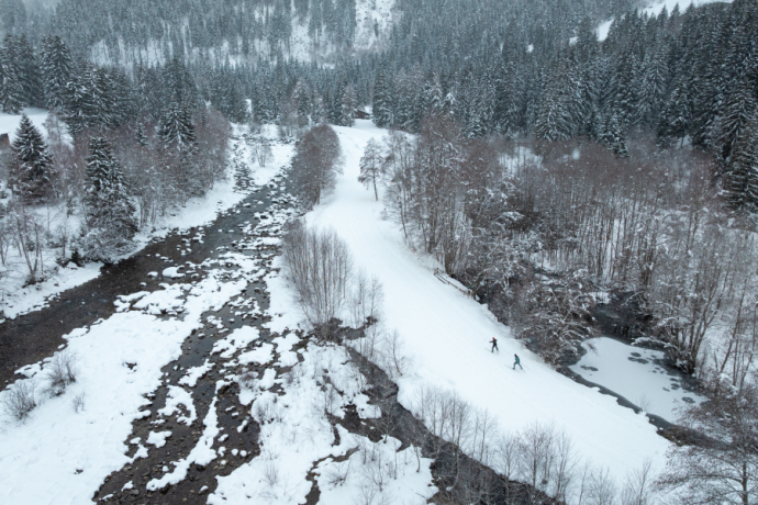 Blick von oben auf Langläufer neben einem Flusslauf im verschneiten Wald bei Disentis