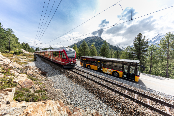 Rhätische Bahn und Postauto begegnen sich vor Bergkulisse in Graubünden