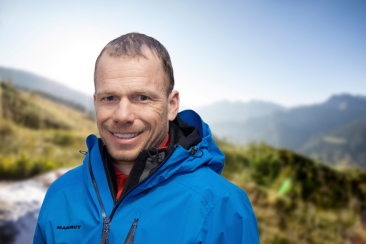 Porträt von Wanderleiter David Baer in blauer Outdoorjacke mit unscharfem Bergpanorama im Hintergrund