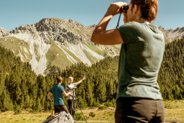 Eine Frau sucht mit einem Fernglas Wildtiere im Schweizerischen Nationalpark, währenddem sich ein Mann und eine Frau im Hintergrund unterhalten
