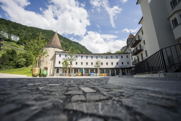 Aussenansicht des Bogentrakt Hostels im Sennhof Chur mit historischem Rundturm heller Fassade und bunter Terrasse vor grünem Berghintergrund