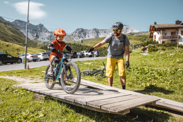 Ein Guide zeigt einem Kind auf einem Mountainbike im Skill Center Arosa, wie es über ein Holzhindernis fahren kann