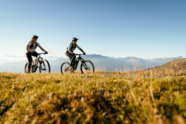 Anita und Caro Gehrig – zwei Mountainbike-Profis – fahren auf einem sonnigen Berghang mit weitem Alpenpanorama im Hintergrund