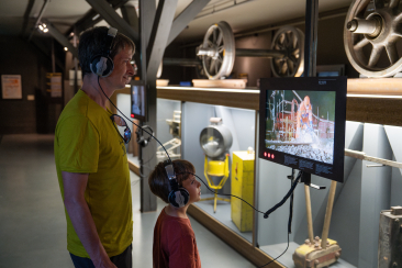 Vater und Sohn mit Kopfhörern vor einem Bildschirm im Bahnmuseum Albula in Bergün, umgeben von historischen Eisenbahnteilen