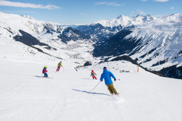Skifahrer*innen fahren bei Sonnenschein eine breite Piste in der SkiArena Andermatt-Sedrun hinunter, im Hintergrund ein verschneites Tal und umliegende Berglandschaft