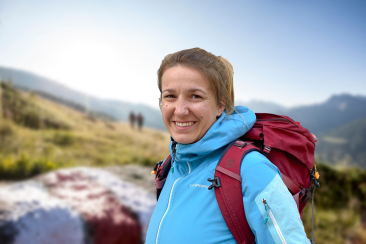 Wanderleiterin Elisabeth Hunziker steht mit rotem Rucksack und blauer Jacke lächelnd auf einem Bergweg vor verschwommenem Alpenhintergrund