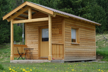 Kleine Holzhuette mit ueberdachter Veranda, Tisch und zwei Stuehlen, auf einer Wiese mit gelben Blumen vor Waldrand.