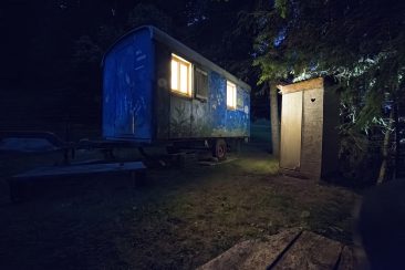 Ein stimmungsvoll beleuchteter, blau bemalter Bauwagen mit warmem Licht aus den Fenstern, daneben ein einfaches Holzplumpsklo mit Herzchen-Ausschnitt inmitten von Bäumen bei Nacht