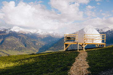 Eine Weisse Bubble-Suite, auf einer Holzplattform mit Stuhl, umgeben von grünen Wiesen, im Hintergrund die schneebedeckten Bündner Alpen unter blauem Himmel.