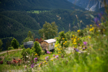 Holzwagen mit rundem Dach auf einem Hang mit bunten Wildblumen, umgeben von Bäumen und mit Blick auf ein bewaldetes Tal