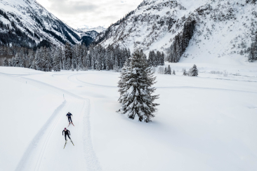 Zwei Langläufer gleiten auf einer verschneiten Loipe durch eine weite Winterlandschaft, umgeben von Bergen und verschneiten Wäldern im Hintergrund.