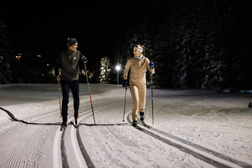 Zwei Langläufer gleiten bei Nacht auf einer beleuchteten Loipe durch einen verschneiten Wald. Der Himmel ist dunkel, und die Scheinwerfer werfen lange Schatten auf den Schnee.
