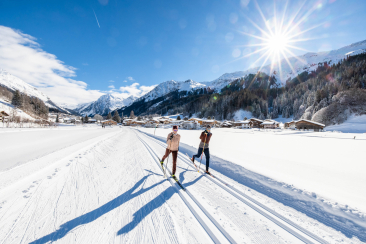 Zwei Langläufer geniessen bei strahlendem Sonnenschein die präparierte Loipe, mit verschneiten Bergen und einem Dorf im Hintergrund.