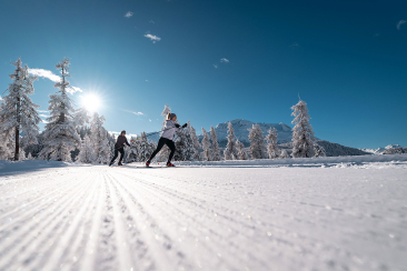 Zwei Langläufer bewegen sich auf einer frisch präparierten Loipe, umgeben von schneebedeckten Bäumen und Bergen im Hintergrund.