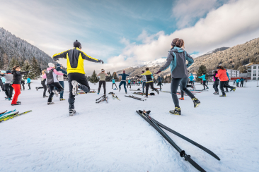 ChatGPT: Gruppe beim Aufwaermen auf Schnee; Langlaufskier liegen daneben, Winterberge und Hauser im Hintergrund.