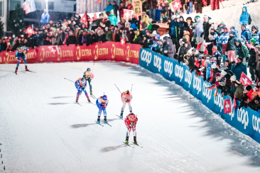 Langlauf-Sprint bei Flutlicht; vier Läufer, dichte Zuschauermenge mit Fahnen entlang der Bande.