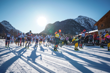 ChatGPT: Start eines Skilanglauf-Massenrennens bei Sonnenschein; viele Laeufer, lange Schatten, Berge und Zuschauer mit Fahnen im Hintergrund.