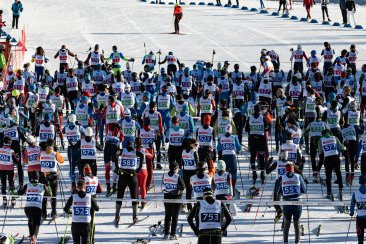 Massenstart eines Langlaufrennens; dichtes Feld mit Startnummern, Skier und Stoecke, Ordner vorn auf der Spur.