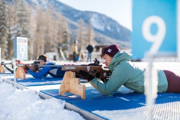 Eine Person liegt beim Biathlontraining auf einer Matte im Schnee und zielt mit einem Gewehr auf eine Schiessscheibe.