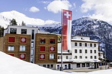 Moderne und traditionelle Gebäude in einer verschneiten Alpenlandschaft mit schneebedeckten Bergen im Hintergrund.