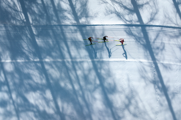 Drei Langläufer gleiten nebeneinander auf einer verschneiten Loipe, aus der Vogelperspektive aufgenommen