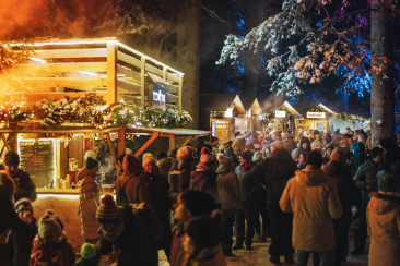 Besucherinnen und Besucher flanieren zwischen beleuchteten Marktständen im winterlichen Zauberwald Lenzerheide