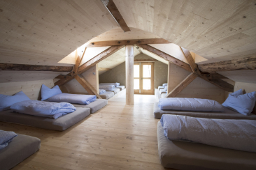 Schlafraum der Gruppenunterkunft Alp Nurdagn im Agrotourismusbetrieb, mit mehreren Matratzenlagern unter dem Dach und hellen Holzelementen