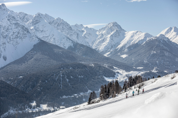 Eine kleine Gruppe von Personen wandert mit Schneeschuhen einen sonnigen Hang im Unterengadin hinauf, umgeben von verschneiten Wäldern und eindrucksvollen Bergen im Hintergrund.