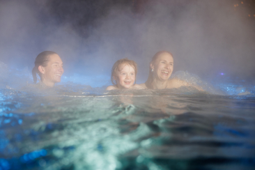 Drei Personen geniessen gemeinsam ein warmes Becken im Familienbad Zernez; sie lachen, während rundherum Dampf über der Wasseroberfläche aufsteigt.
