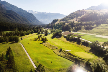 Golfplatz Alvaneu aus der Vogelperspektive bei warmem Abendlicht, grüne Fairways und Bäume vor Bergkulisse.