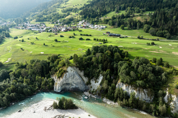 Golfplatz oberhalb des Rheins, grüne Fairways vor Waldhängen und Sagogn im Hintergrund.