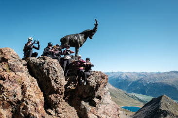 Gruppe von Bikerinnen mit Helmen posiert auf einem Felsgipfel neben einer lebensgrossen Steinbock-Skulptur mit Blick auf die Engadiner Bergwelt